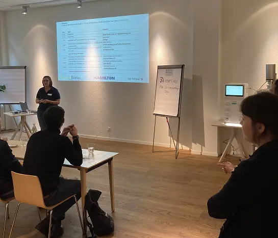 Speaker presenting to seated guests at a Resea Biotec symposium in Bern, with a Hamilton slide on screen